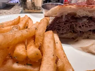 Close-up of a French dip at AJ's Restaurant with thick-cut fries, melted cheese, caramelized onions, and a metal sauce cup behind the sandwich