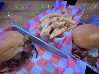 Two smash burgers in paper-lined metal trays with a pile of seasoned fries between them on a wooden table under colorful reflected lights