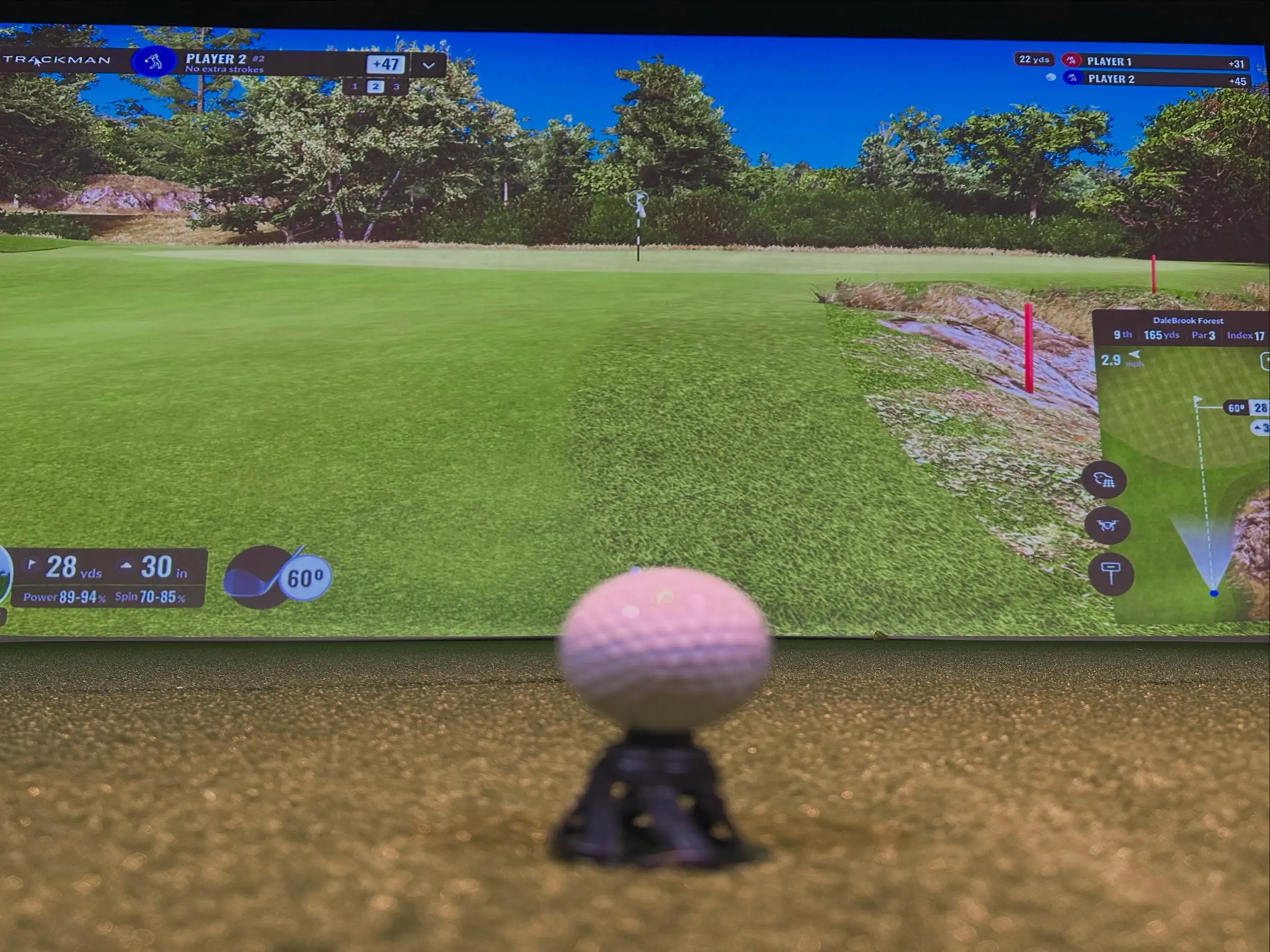 Golf ball on a tee in the foreground of an indoor simulator bay, facing a Trackman virtual green with score overlays and a hole map on screen.