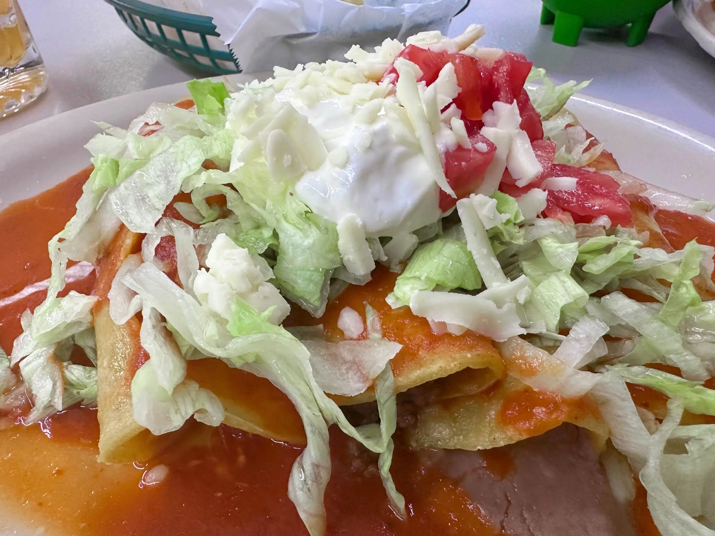Close-up of enchiladas covered in red sauce, shredded lettuce, diced tomato, sour cream, and crumbled white cheese on a white plate