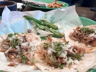 Close-up of carnitas street tacos topped with chopped onion and cilantro beside grilled jalapeños and onions, with arroz con pollo in the background