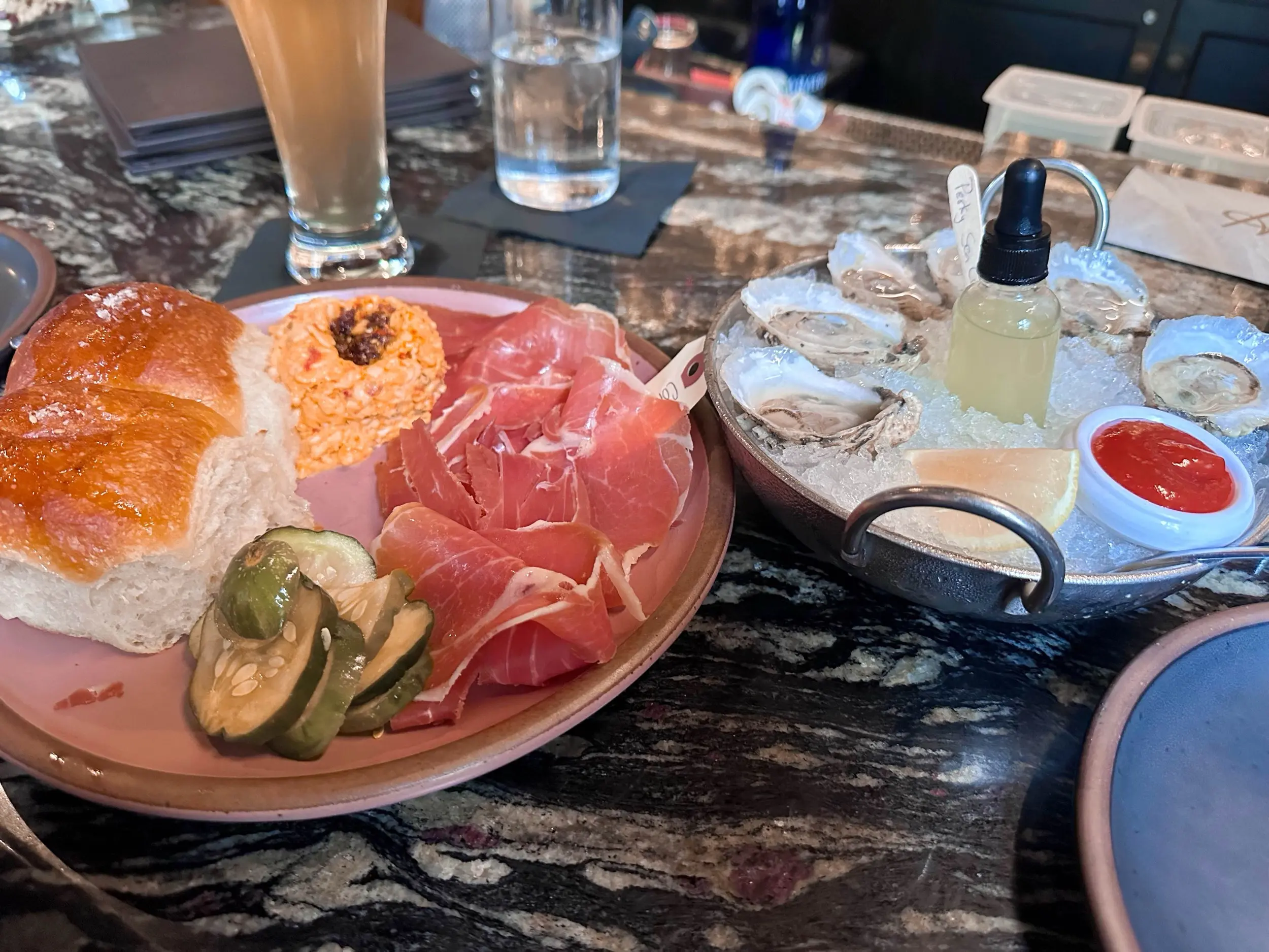 A bar-top spread with oysters on ice beside rolls, sliced ham, a pimento spread, pickles, and drinks at Husk Charleston
