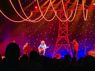 Jesse Welles performs with an acoustic guitar at The Pinnacle, flanked by a pianist and a guitarist, beneath a glowing red electricity pylon and a starry backdrop as silhouetted fans watch.