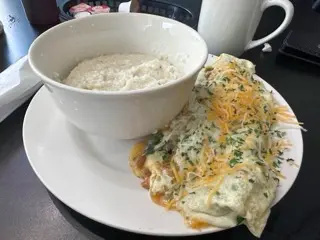 A large bowl of grits beside a cheese-covered omelet on a white plate, with a mug of coffee on the table at Lil Jazzie's