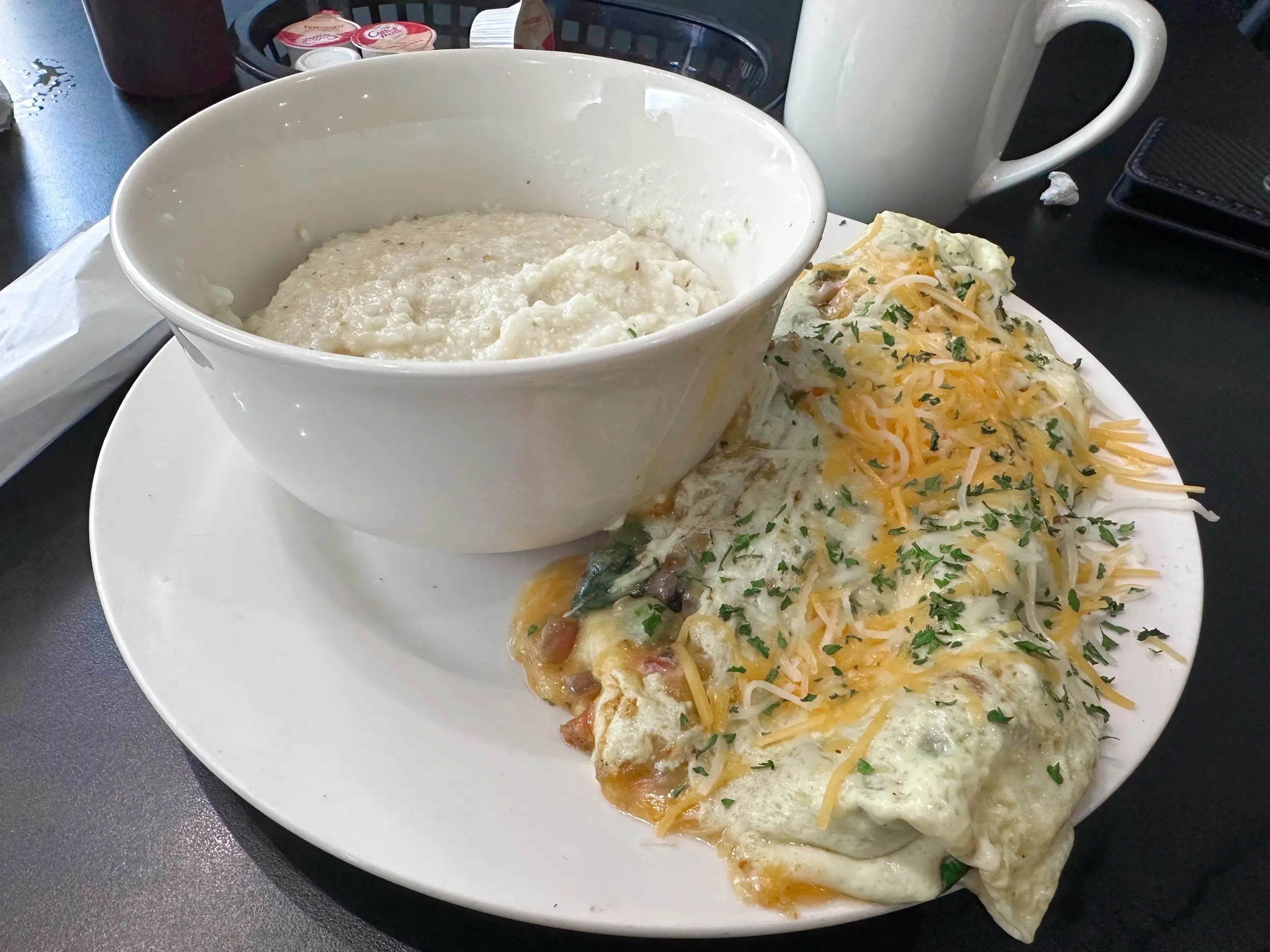 A large bowl of grits beside a cheese-covered omelet on a white plate, with a mug of coffee on the table at Lil Jazzie's