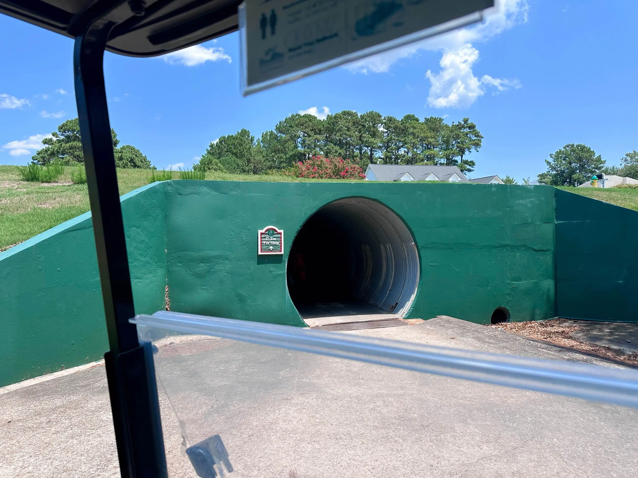 View from a golf cart at Myrtlewood Golf Club facing a wide green tunnel under a path, with bright summer sky, trees, and low buildings beyond the embankment.