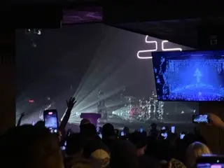 A dark concert crowd holding up phones beneath blue and purple stage lights, with a performer projected on a large screen and an illuminated geometric symbol overhead