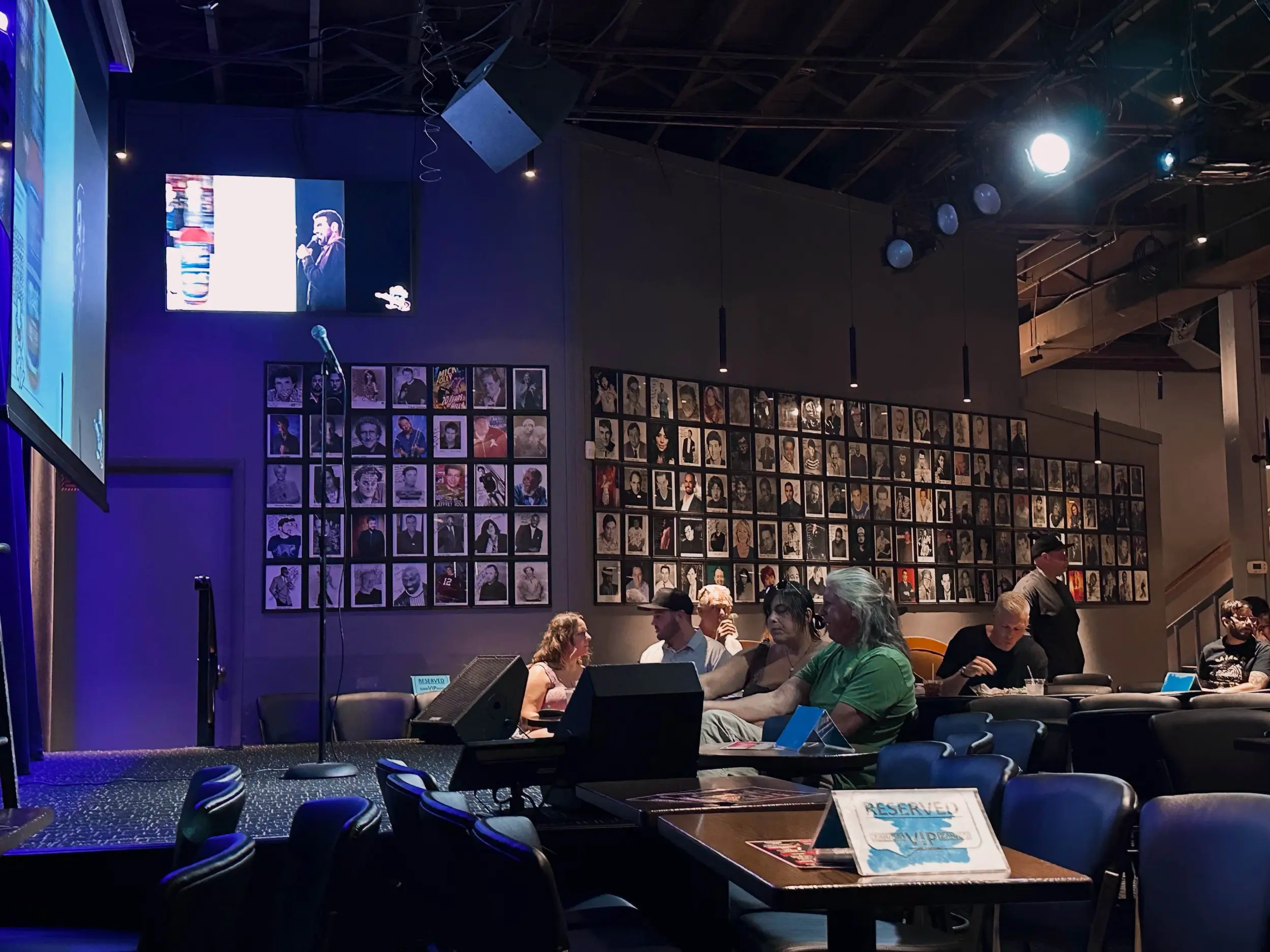The empty microphone and low stage at Zanies Nashville before the show, with reserved tables, seated guests, blue-purple lighting, and walls covered in framed comedian portraits