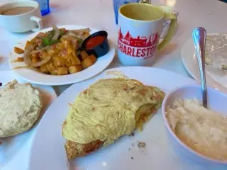 Ham, bacon, and cheese omelet with grits and a biscuit at Toast! on Meeting, with coffee, an Americano mug, and potatoes with peppers and onions on the table