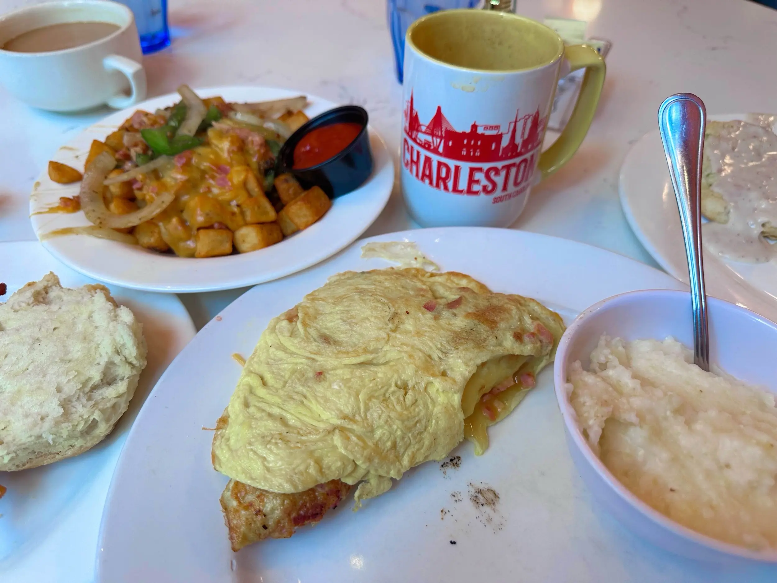 Ham, bacon, and cheese omelet with grits and a biscuit at Toast! on Meeting, with coffee, an Americano mug, and potatoes with peppers and onions on the table