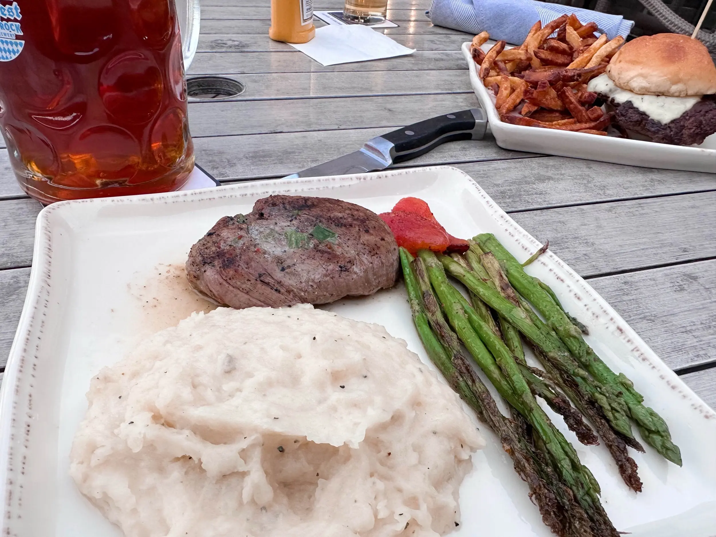 Filet mignon with mashed potatoes, asparagus, and roasted tomatoes in the foreground, with an amber beer and elk burger with fries across the patio table