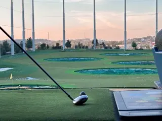 Driver resting beside a golf ball on a Topgolf bay mat with the Nashville outfield targets, tall netting poles, and a soft pink evening sky in the distance.