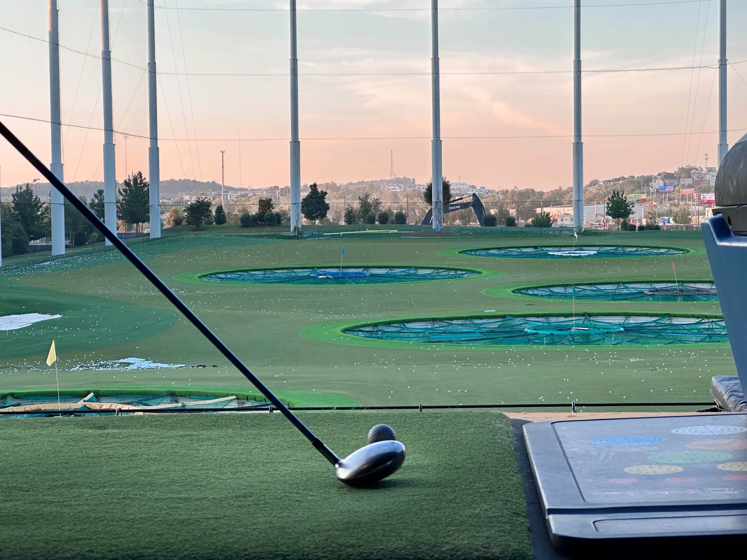 Driver resting beside a golf ball on a Topgolf bay mat with the Nashville outfield targets, tall netting poles, and a soft pink evening sky in the distance.