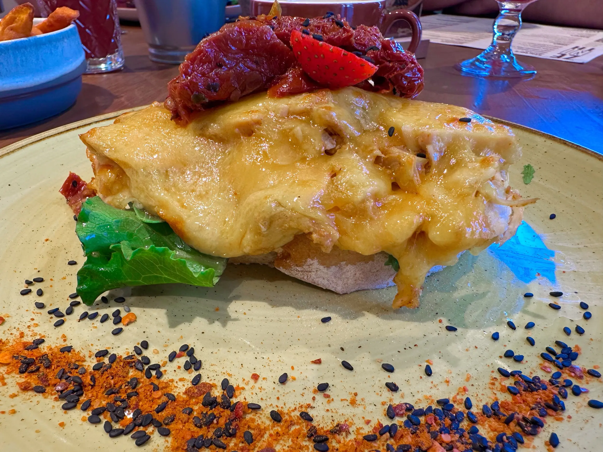 A chicken toast at Zenith Caffè in Matosinhos covered in melted cheese and topped with dried tomato, strawberry, and black sesame on a tan plate